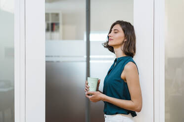 Junge Geschäftsfrau mit geschlossenen Augen, die eine Einweg-Kaffeetasse hält, während sie an der Tür im Büro lehnt - DIGF15497