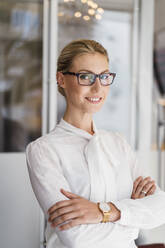 Lächelnde blonde Geschäftsfrau mit Brille steht mit verschränkten Armen im Büro - DIGF15501