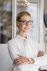 Smiling blond businesswoman wearing eyeglasses standing with arms crossed in office - DIGF15501