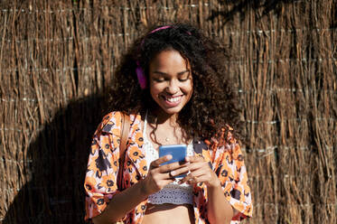 Smiling woman using smart phone in front of straw wall on sunny day - KIJF03877