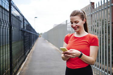 Lächelnde, fitte Frau, die auf dem Gehweg eine Textnachricht auf ihrem Smartphone schreibt - ASGF00338