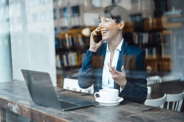 Mature female entrepreneur with laptop talking on mobile phone at cafe - JOSEF04589