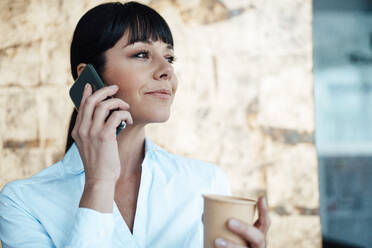 Female entrepreneur holding coffee cup while talking on mobile phone - JOSEF04617