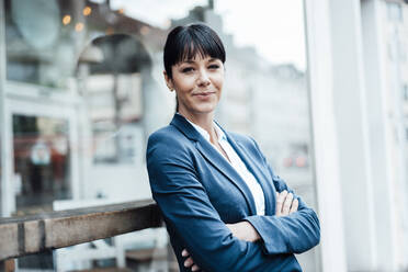 Smiling female entrepreneur with arms crossed leaning on cafe window - JOSEF04624