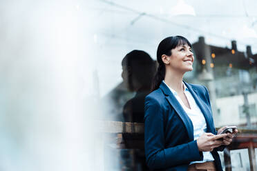 Thoughtful businesswoman holding mobile phone while leaning on cafe window - JOSEF04626