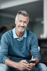 Smiling businessman holding mobile phone while sitting at cafe - JOSEF04718