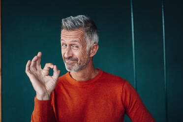 Mature man gesturing OK sign in front of wall - JOSEF04768