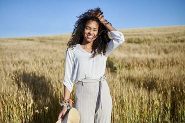 Lächelnde junge Frau steht mit der Hand im Haar in einem Weizenfeld an einem sonnigen Tag - KIJF03895