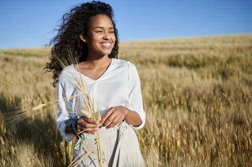 Lächelnde junge Frau hält Weizenernte auf einem Feld an einem sonnigen Tag - KIJF03900