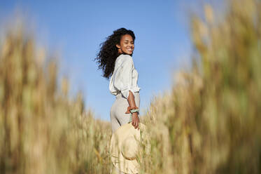 Lächelnde, lockig behaarte Frau mit Hut in einem Weizenfeld an einem sonnigen Tag - KIJF03912