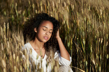Schöne junge Frau mit geschlossenen Augen sitzt in Weizenfeld während sonnigen Tag - KIJF03914