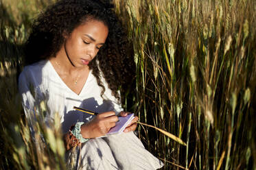 Young woman with curly hair writing in notepad while sitting at wheat field during sunny day - KIJF03916