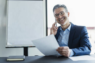 Lächelnder Geschäftsmann mit Brille, der im Büro mit einem Mobiltelefon spricht und wegschaut - DIGF15506