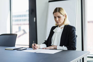 Blond businesswoman writing while sitting at desk in office - DIGF15508