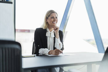 Lächelnde Unternehmerin, die mit der Hand am Kinn am Schreibtisch sitzt und wegschaut - DIGF15512