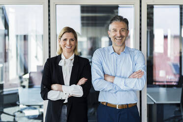 Smiling businessman and businesswoman standing with arms crossed at doorway in office - DIGF15520