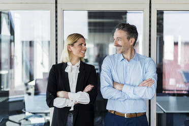 Smiling male and female professionals with arms crossed looking at each other while standing at doorway in office - DIGF15522