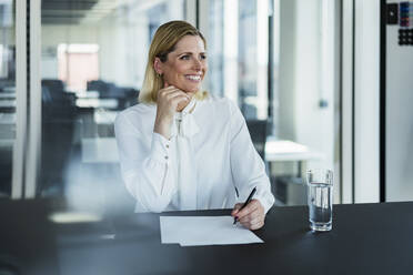 Smiling female entrepreneur sitting with hand on chin at desk looking away - DIGF15531