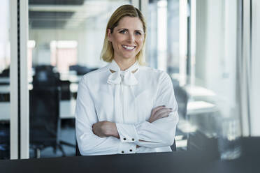 Smiling female professional sitting with arms crossed at desk - DIGF15532