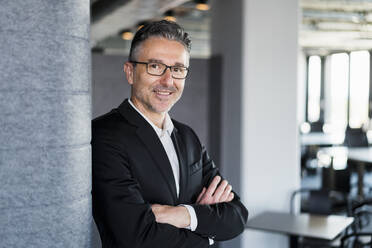 Smiling male entrepreneur standing with arms crossed by wall in office - DIGF15550