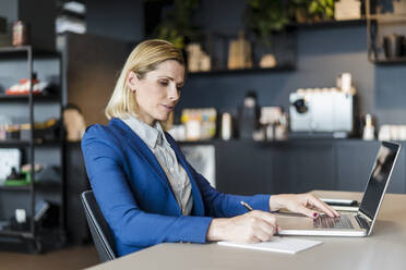 Blond businesswoman writing while sitting with laptop at desk in office - DIGF15559