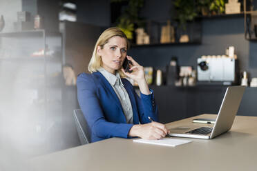 Blonde Frau spricht mit ihrem Smartphone, während sie am Schreibtisch im Büro sitzt - DIGF15561