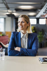 Smiling female professional sitting with arms crossed at desk in office - DIGF15564