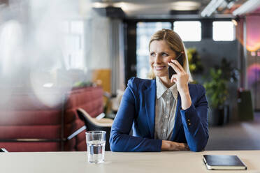 Lächelnde blonde Geschäftsfrau, die am Schreibtisch sitzend mit ihrem Handy telefoniert und wegschaut - DIGF15565