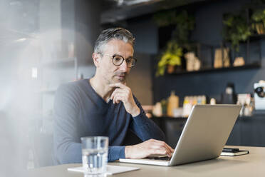 Männlicher Berufstätiger sitzend mit Hand am Kinn und Laptop am Schreibtisch im Büro - DIGF15573