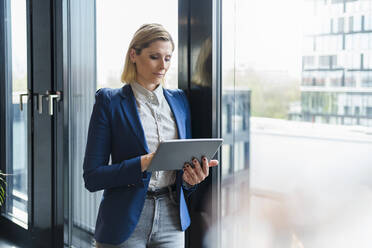 Geschäftsfrau, die ein digitales Tablet benutzt, während sie an einem Glasfenster im Büro steht - DIGF15578