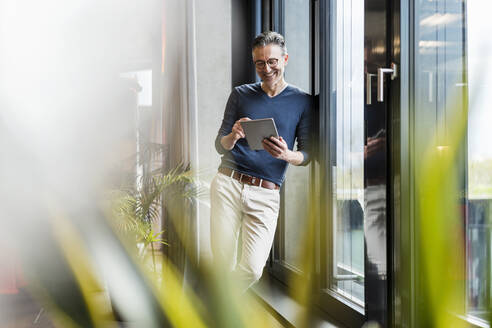 Fröhlicher Geschäftsmann, der ein digitales Tablet benutzt und sich an ein Glasfenster im Büro lehnt - DIGF15580