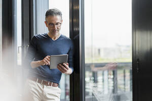 Männlicher Unternehmer, der ein digitales Tablet benutzt und sich an ein Glasfenster im Büro lehnt - DIGF15581