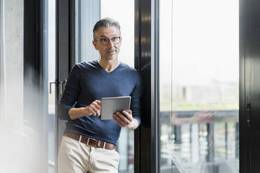 Mature male professional with digital tablet leaning on glass window while looking away - DIGF15582