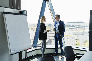 Female professional discussing with businessman while standing by glass window in board room - DIGF15590