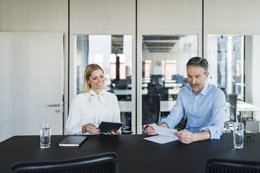 Smiling businesswoman looking at male colleague reading documents while sitting at desk in office - DIGF15619