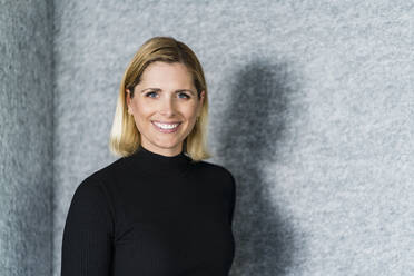 Smiling female entrepreneur with blond hair standing in front of wall - DIGF15630