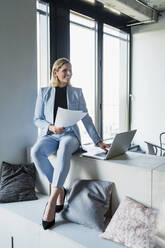 Lächelnde Geschäftsfrau mit Dokumenten und Laptop auf Möbeln im Büro sitzend - DIGF15636