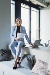 Smiling businesswoman with documents and laptop sitting on furniture in office - DIGF15636