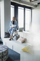 Smiling female entrepreneur with documents and laptop sitting on furniture in office looking away - DIGF15637