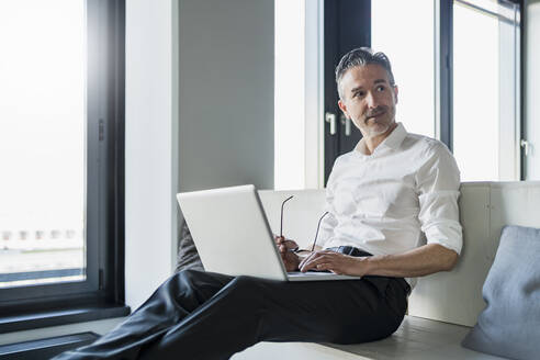 Männlicher Berufstätiger sitzt mit Laptop auf Möbeln im Büro und schaut weg - DIGF15645