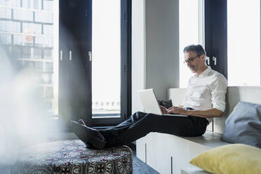 Mature businessman using laptop sitting on furniture while working in office - DIGF15646