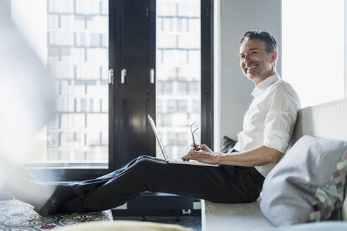 Smiling male entrepreneur sitting with laptop on seat in office while looking away - DIGF15647