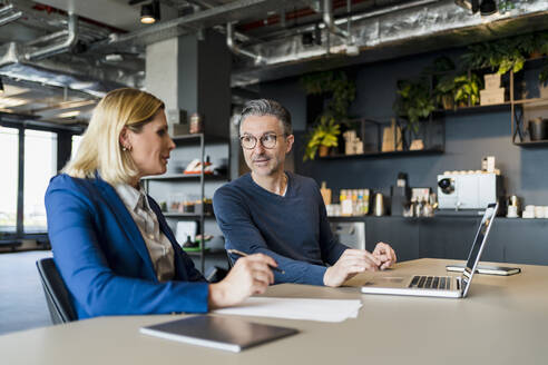 Männlicher Unternehmer im Gespräch mit einer weiblichen Fachkraft, die mit einem Laptop in einem kreativen Büro sitzt - DIGF15652