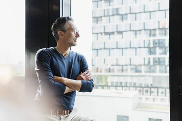 Mature businessman standing with arms crossed looking through window - DIGF15658