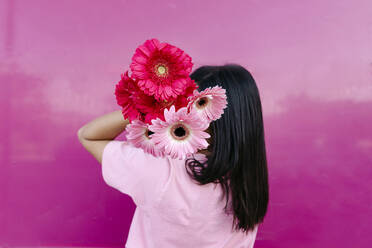 Junge Frau versteckt Gesicht mit rosa Gerbera-Gänseblümchen an der Wand - TCEF01894