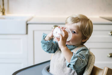 Blonder Junge trinkt Milch, während er auf einem Hochstuhl in der Küche zu Hause sitzt - XLGF01925