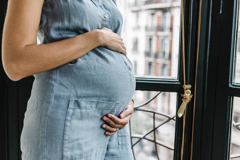 Schwangere Frau mit Hand auf dem Bauch, die zu Hause am Fenster steht - XLGF01927