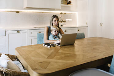 Geschäftsfrau mit Sohn arbeitet am Laptop am Esstisch in der Küche - XLGF01936