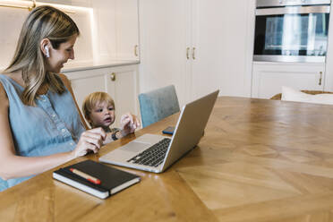 Smiling female freelancer looking at curious son in front of laptop at dining table in kitchen - XLGF01943