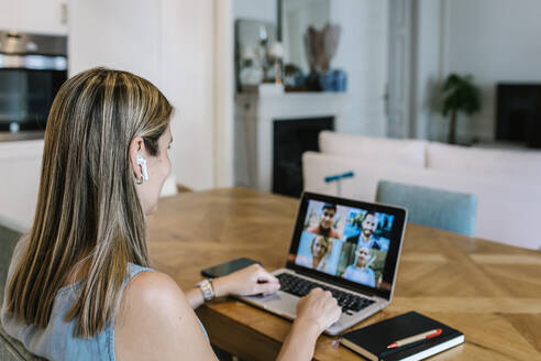 Female freelance worker wearing wireless earphones talking on video conference through laptop at home - XLGF01944